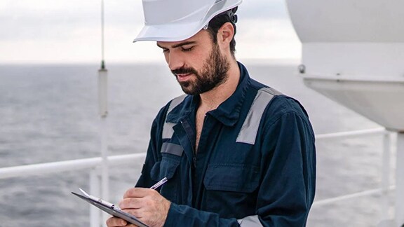Person standing on a ship’s deck wearing protective work clothing and a hard hat, holding a tablet with the ocean visible in the background.